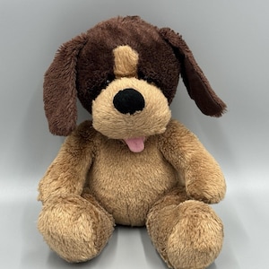 May include: A plush dog toy with brown and tan fur. The dog has a brown head and ears, a tan snout, and a pink tongue sticking out. The toy is sitting upright with its paws forward, and has a black nose.