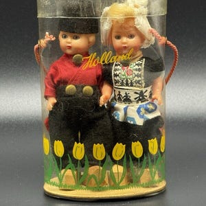 May include: A pair of Dutch dolls in traditional attire, enclosed in a clear plastic container. The dolls are dressed in black and red outfits, with the word "Holland" printed on the container. The container is decorated with yellow tulips.