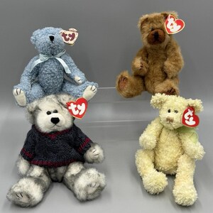 May include: Four plush teddy bears in various colors: blue, brown, gray, and yellow. Each bear has a heart-shaped tag attached. The bears are sitting on a gray surface.