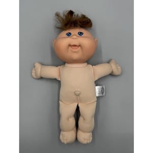 Puede incluir: Una muñeca Cabbage Patch Kids con cuerpo de tela suave de tono claro, cabeza redonda de vinilo y cabello castaño peinado en un moño desordenado. La muñeca tiene ojos azules brillantes, una boca pequeña abierta y un ombligo. Etiqueta blanca.