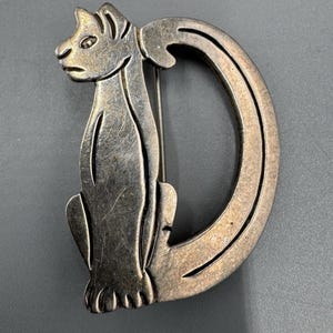 May include: A silver-toned metal brooch shaped like a cat within the letter "D". The cat has etched details, and the brooch has a pin for fastening. The brooch is set against a grey background. The brooch is approximately 5 cm in length.