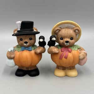 May include: Two ceramic bear figurines. One bear wears a black top hat and holds a black bird and a white bag. The other bear wears a yellow bonnet and holds a black bird and a pink bag. Both bears are dressed in orange pumpkin costumes.
