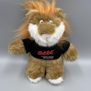 May include: A plush lion toy with brown fur, a white muzzle, and a tuft of orange hair on its head. The lion wears a black t-shirt with the red text D.A.R.E. and the white text TO RESIST DRUGS AND VIOLENCE printed on the front of the shirt.