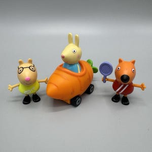 Op de afbeelding: Drie plastic speelfiguren waaronder een lichtgeel dier met een bril en een geel shirt, een lichtgeel konijn in een oranje wortelvormig voertuig met zwarte wielen, en een oranje dier dat een paars racket vasthoudt.