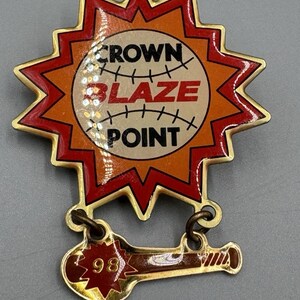 Könnte beinhalten: Eine goldfarbene Anstecknadel mit Baseball-Motiv. Die Anstecknadel zeigt eine rote und orangefarbene Sternform, die einen Baseball umgibt, mit den Worten "CROWN BLAZE POINT". Ein kleineres, angebrachtes Element zeigt "98".