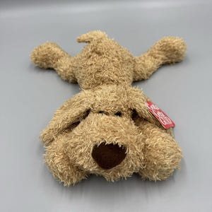 May include: A plush, light brown dog toy with floppy ears and a dark brown nose. The dog is lying on its back with its arms and legs outstretched. A red tag is attached to the toy.