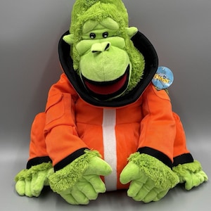 May include: A plush green gorilla wearing an orange jumpsuit with black trim and a white stripe down the front. The gorilla has a black collar and green fur on its head and hands. A tag is attached to the gorilla.