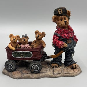 Può includere: Una statuetta in resina di un orso bruno che indossa una camicia a quadri rossi, salopette scura e un berretto con la lettera B, che traina un carro rosso con la scritta BEAR EXPRESS. Nel carro siedono tre orsetti, uno con un vestito rosa.