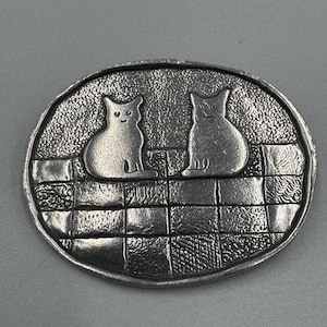 Könnte beinhalten: Eine ovale Gürtelschnalle aus silberfarbenem Metall mit einem Reliefdesign von zwei Katzen, die nebeneinander auf einer texturierten, gitterartigen Oberfläche sitzen. Der Hintergrund hinter den Katzen hat eine körnige Oberfläche.