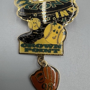 Könnte beinhalten: Baseball-Pin mit den Worten "ZONE IN" und "CROWN POINT" in grünen und goldenen Buchstaben. Der Pin zeigt Basebälle mit lächelnden Gesichtern und einen Baseballhandschuh mit der Nummer "98".