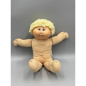 Puede incluir: Una muñeca con cara redonda de tela color melocotón, ojos azul brillante y nariz pequeña. La muñeca tiene el pelo corto de lana rizada de color amarillo pálido y un cuerpo de tela suave de color claro con brazos y piernas articulados.