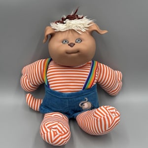 Puede incluir: Una muñeca de peluche con cara de vinilo color canela, nariz marrón y cabello de hilo blanco y marrón. La muñeca viste una camiseta de manga larga a rayas naranjas y blancas, un peto de pana azul con tirantes arcoíris y un parche de elefante.
