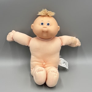 May include: A soft-bodied doll with a round, peach-toned vinyl head and limbs. The doll features blue eyes, a small nose, a closed mouth, and a tuft of blonde yarn hair on top of its head. It has a fabric torso and a white care tag attached to its side.