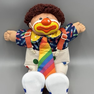 Puede incluir: Una muñeca de peluche con cabello de lana marrón y una máscara de payaso de plástico con nariz roja y boca amarilla. La muñeca viste una camisa azul marino con estampado de crayones coloridos, overol blanco con lunares verdes y rojos, tirantes naranjas y una corbata de rayas arcoíris, terminada con zapatos de plástico blanco.
