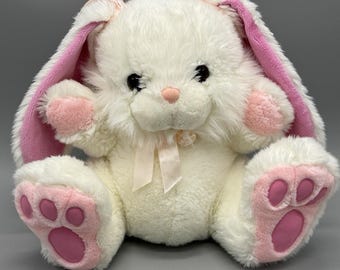 May include: A plush stuffed rabbit toy with white fur, featuring long ears with pink inner lining, a small pink nose, and dark eyes. The rabbit has pink paw pads on its feet and hands, and a light pink ribbon tied around its neck.