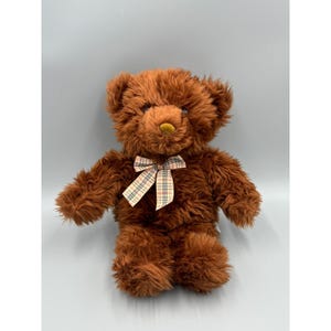 May include: A plush teddy bear with shaggy, medium-brown fur, dark eyes, and a small, rounded nose. The bear features a decorative ribbon tied around its neck, patterned with a classic beige, black, and red plaid design against a soft texture.
