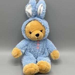 May include: A plush teddy bear wearing a light blue bunny costume with a hood and pink button details. The bear has tan fur on its face, paws, and feet. The bunny ears are light blue with white inner ears. The toy is on a gray background.