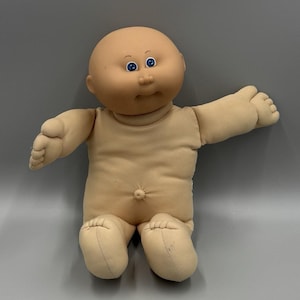 Pode incluir: Uma boneca careca com corpo de tecido bege claro, cabeça redonda de vinil e olhos azul-brilhantes. A boneca apresenta um umbigo proeminente, braços estendidos e pernas em posição sentada, tudo num tom de pele claro e neutro.