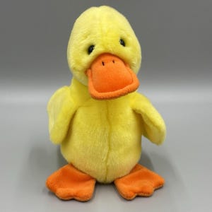 May include: A plush yellow duck with an orange beak and feet. The stuffed animal has small black eyes and is standing upright. The duck is made of soft, fuzzy material and is a classic children's toy.
