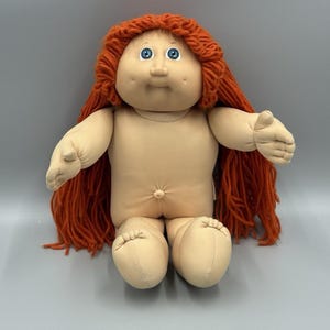 Puede incluir: Una muñeca de cuerpo blando con piel clara, ojos azul brillante y cabello largo de lana naranja. La muñeca presenta una cara redonda con hoyuelos, una nariz pequeña y una hendidura de ombligo en el torso. Los brazos están extendidos y las piernas dobladas.