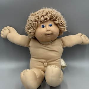 May include: A vintage Cabbage Patch Kid doll with a tan fabric body, arms outstretched, and seated. The doll has a round face, blue eyes, and light brown yarn hair. The doll is wearing a simple tan top.