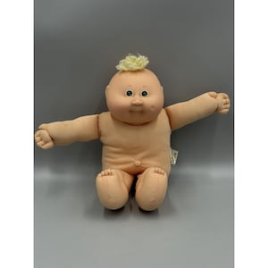 Peut inclure: Une poupée Cabbage Patch Kids avec un corps en tissu souple, un teint clair et une tête ronde. La poupée présente une petite touffe de cheveux en laine blonde sur le dessus, des yeux verts, un petit nez et une fossette sur la joue gauche.