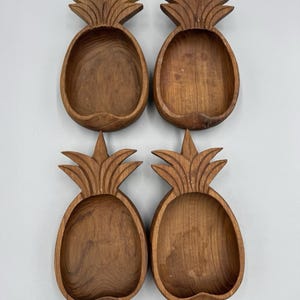 Puede incluir: Cuatro cuencos de madera tallada con forma de piña, dispuestos en una cuadrícula de dos por dos. Cada cuenco presenta una veta de madera marrón natural, un interior liso y un patrón decorativo de hojas talladas en la parte superior.