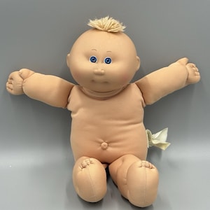 Op de afbeelding: Een zachte pop met een rond, perzikkleurig stoffen hoofd en lichaam, voorzien van helderblauwe ogen, een kleine neus en een plukje lichtblond garen als haar bovenop. De pop heeft een navel en is afgebeeld met de armen wijd gespreid.