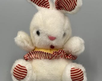 May include: A plush white rabbit toy with soft fur, featuring red and white striped fabric on the inner ears, the soles of the feet, and a ruffled collar. The rabbit has a small pink nose, a dark red mouth, and black eyes, with a yellow shirt.
