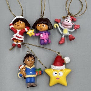 May include: A set of five hanging ornaments featuring characters from Dora the Explorer. The figures include Dora in a Santa outfit, Dora in winter gear with a star, a monkey in a red hat, a boy holding a gift and small animal, and a yellow star in a Santa hat.