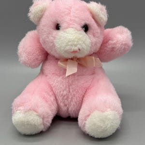 Può includere: Un orsacchiotto di peluche con morbida pelliccia rosa e accenti bianchi a contrasto su orecchie, muso e piedi. L'orso ha occhi scuri e rotondi, un piccolo naso rosa e un nastro di raso rosa chiaro legato attorno al collo, seduto in posizione eretta.