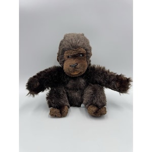 May include: A plush gorilla toy featuring dark brown, shaggy synthetic fur, a smooth brown face, black plastic eyes, and a small black nose. The stuffed animal is depicted in a seated position with arms extended outward and legs tucked forward.