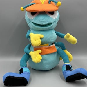 May include: A plush toy bug with a light blue body, orange hat, and yellow hands and accents. The hat features a lightbulb graphic. The bug has blue legs and feet with black soles. The toy is designed with a friendly, cartoonish appearance.
