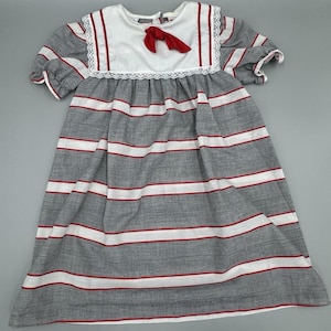 Könnte beinhalten: Ein grau-weiß gestreiftes Kleid mit roten Akzenten. Das Kleid hat kurze Ärmel, einen weißen Kragen mit roter Bordüre und eine kleine rote Schleife. Das Kleid hat einen klassischen, Vintage-Stil.