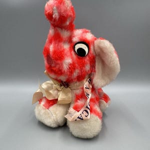 Op de afbeelding: Een vintage pluche konijn met rood en wit geruit bont. Het heeft grote zwarte ogen, roze gevoerde oren en een crèmekleurige strik. Een lint met het woord "AMERICA" is bevestigd. Het speelgoed is een nostalgisch verzamelobject.