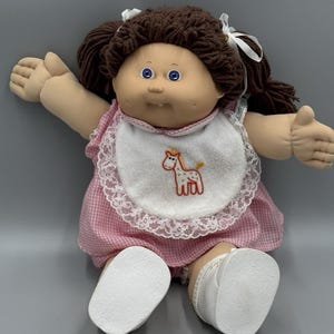 May include: A doll with brown yarn hair tied in pigtails with white ribbons, blue eyes, and a single visible tooth. The doll wears a pink and white gingham dress and a white bib featuring an embroidered giraffe, paired with white shoes.