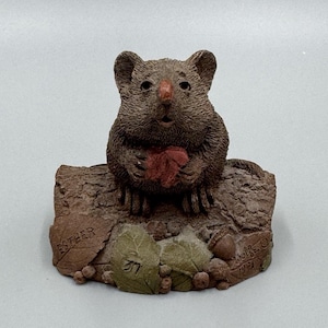 May include: A brown mouse figurine sitting on a textured log base. The mouse holds a small red leaf. The base features sculpted leaves, acorns, and berries, with the name ESTHER inscribed on a leaf and the year 1991 visible on the right side.