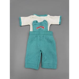 Peut inclure: Un t-shirt en tricot blanc à manches courtes avec des bordures bleu sarcelle, associé à une salopette en tricot bleu sarcelle. La salopette présente un logo rouge brodé sur la poitrine indiquant Cabbage Patch Kids en maille texturée.