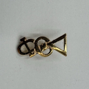 Peut inclure: Une broche en métal doré composée de trois lettres grecques entrelacées. Le motif comprend la lettre Phi, suivie d'une lettre circulaire, et se termine par une forme triangulaire Delta. L'objet présente une finition métallique polie.