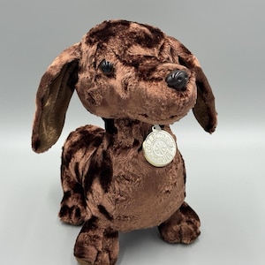 May include: A plush brown dachshund dog with a soft, textured appearance. The stuffed animal has floppy ears, a black nose, and a silver tag around its neck. The tag reads "Collectors Edition".