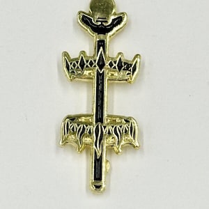 May include: A gold-colored metal pin with a black enamel design. The pin features a cross-like shape with ornate details, including crown-like elements and a circular top. The design is symmetrical and detailed, with a shiny finish.
