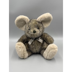 May include: A plush mouse toy with dark brown and gray mottled fur, large beige ears, and beige paws. The stuffed animal features a small black nose and a smooth gray ribbon tied in a bow around its neck, sitting in an upright position.