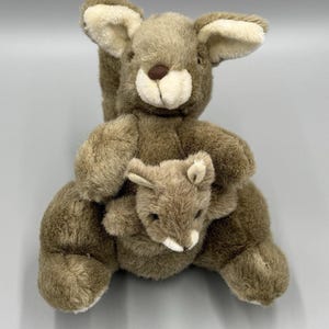 May include: A plush kangaroo toy in shades of brown and beige. The larger kangaroo has a smaller joey in its pouch. The toy has soft, fluffy fur and detailed facial features, including a brown nose and white accents.