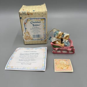 May include: A Cherished Teddies hanging ornament featuring two teddy bears in a pink sleigh, accompanied by a cardboard box, a certificate of authenticity, and a small informational card. The bears wear blue hats and scarves, with the text Our 1st Christmas.