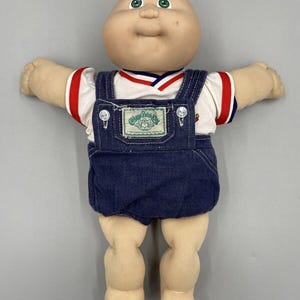 Puede incluir: Una muñeca Cabbage Patch Kids vintage con cabeza calva, ojos verdes y expresión sonriente. La muñeca viste una camisa blanca con ribetes rojos y azules, y un peto vaquero azul. El peto tiene un parche que dice "Cabbage Patch Kids".