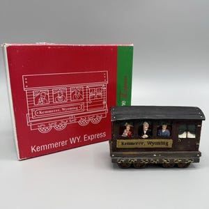 May include: A miniature train car with the text "Kemmerer WY. Express" on the side and box. The train car is brown with small figures inside. The box is red with a green stripe.