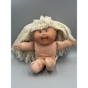 Pode incluir: Uma boneca de corpo macio com um rosto redondo cor de pêssego, olhos verdes e uma pequena boca aberta mostrando um único dente. A boneca apresenta cabelos longos e grossos de lã cor de creme com franja, braços estendidos e pernas dobradas.