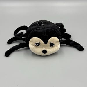 May include: A plush black spider toy with eight curved legs. The spider has a cream-colored face with large, round eyes and a small black nose. A metal ring is attached to the top of the spider's head.