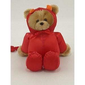 Op de afbeelding: Een pluche teddybeer met lichtbruine vacht, gekleed in een felrood, satijnachtig kostuum met capuchon. De capuchon heeft twee kleine oranje hoorntjes bovenop en wordt onder de kin gestrikt met een rood lint. De beer heeft donkere ogen en een donkere neus.