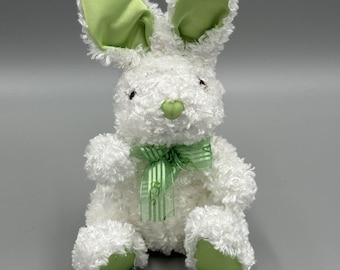 May include: A plush stuffed rabbit toy with white curly fur, featuring light green fabric accents on the inner ears, nose, and foot pads. The rabbit is adorned with a sheer, light green striped ribbon tied in a bow around its neck.